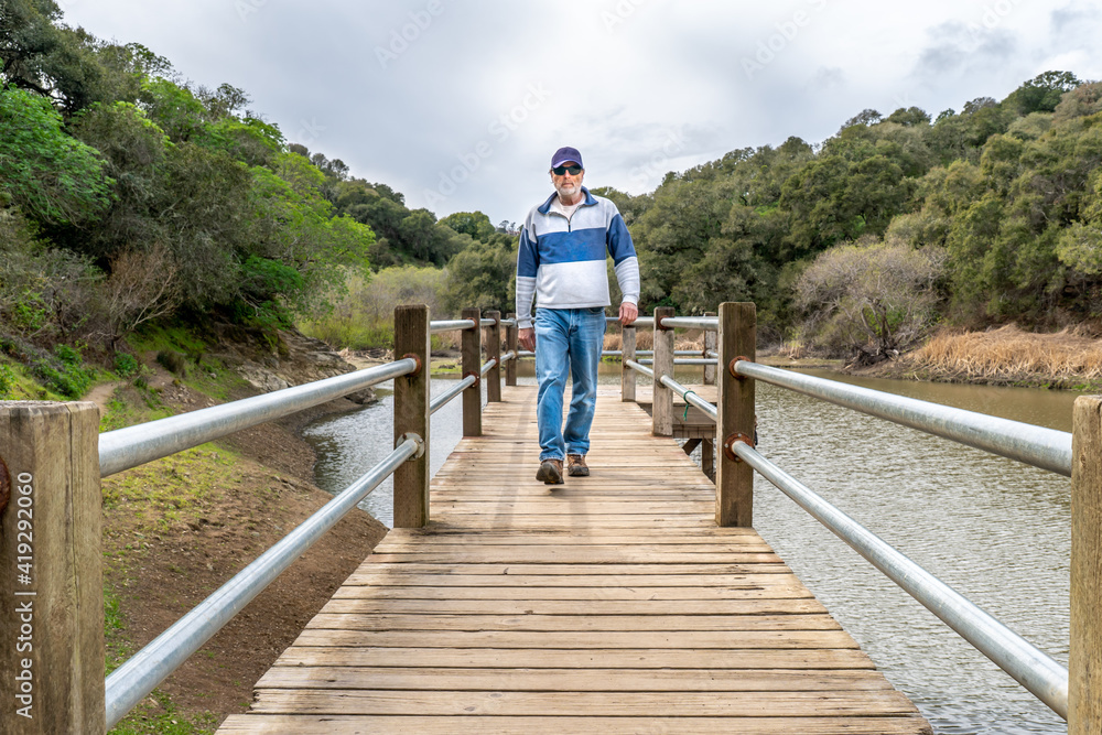A man, visitor, hiker walking towards on a pier or a wharf over a small lake with tree covered trees on both sides and cloudy sky overhead, Waterdog Lake, Belmont, California