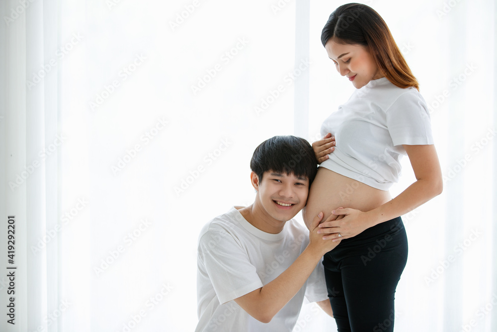 young lovely Asian husband and wife stay together in the living room. A pregnant woman standing and looking at her belly while a man sitting and listening to a baby in her belly with love copy space young lovely Asian husband and wife stay together in the living room. A pregnant woman standing and looking at her belly while a man sitting and listening to a baby in her belly with love copy space