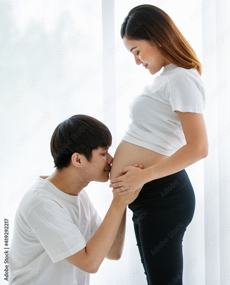 portrait of a very lovely young Asian couple. A husband sitting and kissing a wife's belly while a pregnant woman standing smiling and looking at a man with much love portrait of a very lovely young Asian couple. A husband sitting and kissing a wife's belly while a pregnant woman standing smiling and looking at a man with much love