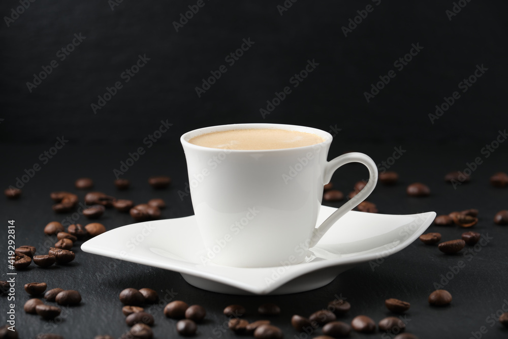 Fototapeta premium White cup of coffee on a black background. Close-up side view. Coffee beans on a black table.