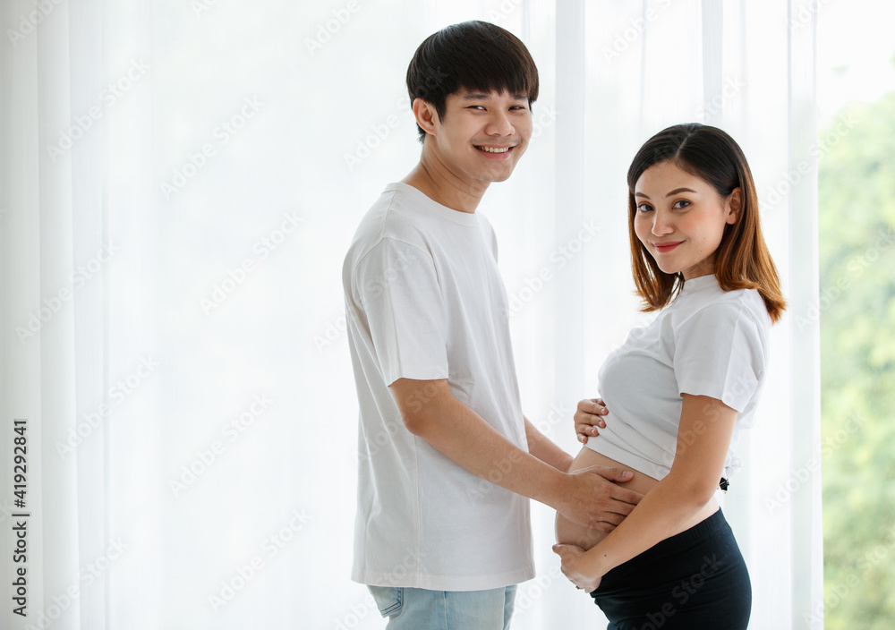 Fototapeta premium A lovely young Asian couple husband and wife standing together smiling and touching a pregnant woman's belly with love and care. Happy and healthy family concept.