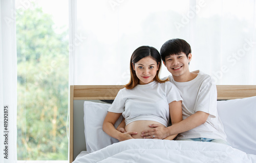 portrait of a good-looking young Asian man and woman wearing a white nightdress sitting on a bed together. They are smiling happily and touching the belly of a pregnant mother