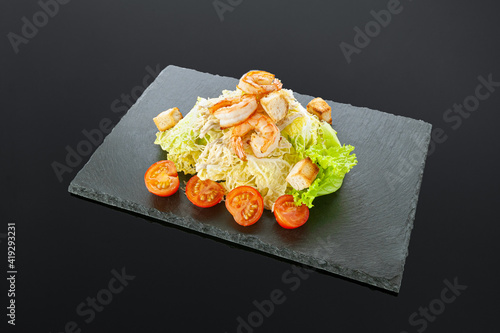 Caesar salad with shrimps. On a black slate board. Salad ingredients: cherry tomatoes, salad mix, croutons, parmesan cheese, sauce. Dark background. Isolated. Close-up.