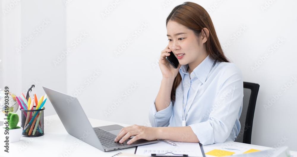 Asian female employee talking on the phone and working in a laptop with a smiling face, Office staff work and telephone conversations, Business conversation over the phone. Asian female employee talking on the phone and working in a laptop with a smiling face, Office staff work and telephone conversations, Business conversation over the phone.