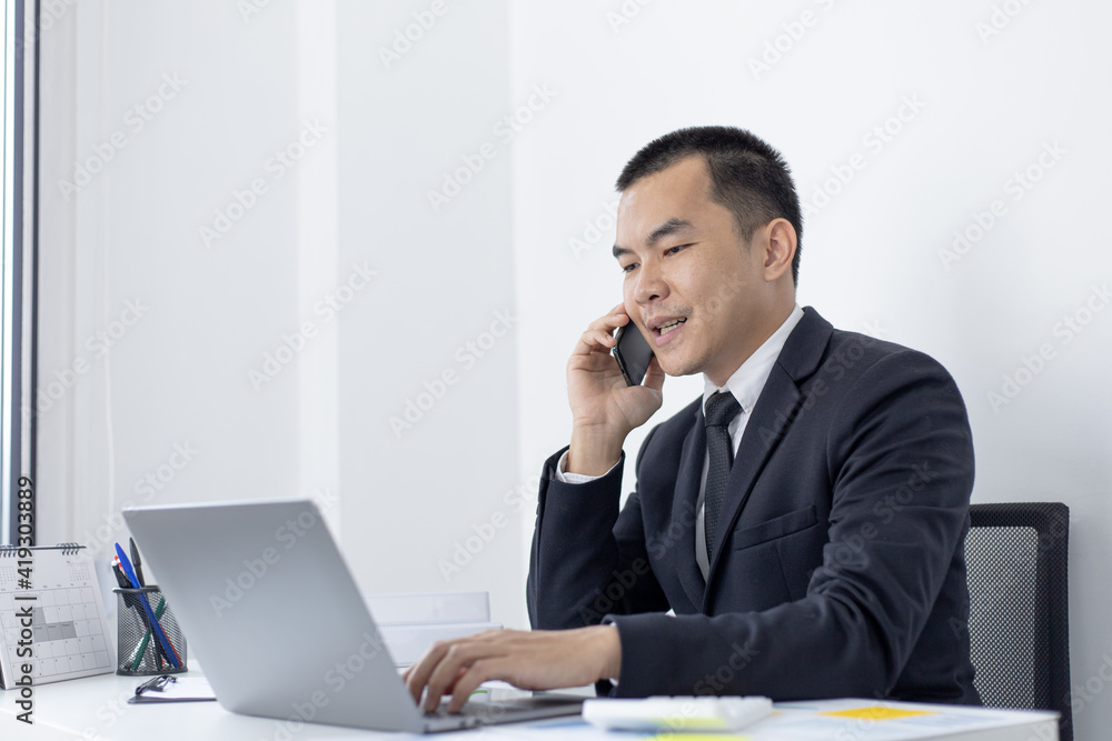 Asian man employee talking on the phone and working in a laptop with a smiling face, Office staff work and telephone conversations, Business conversation over the phone, handsome male in a black suit. Asian man employee talking on the phone and working in a laptop with a smiling face, Office staff work and telephone conversations, Business conversation over the phone, handsome male in a black suit.