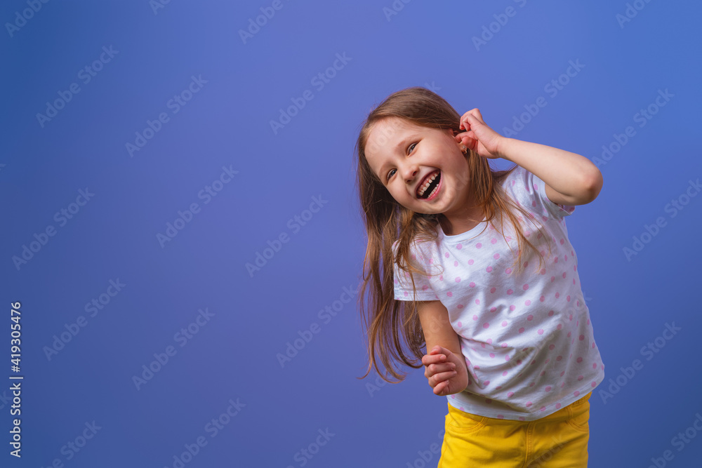 Fototapeta premium Little girl 5 years old with blonde hair in a T-shirt with colored polka dots standing happily smiling on purple background. Close-up. the model is having fun and feels overwhelmed, amazed.