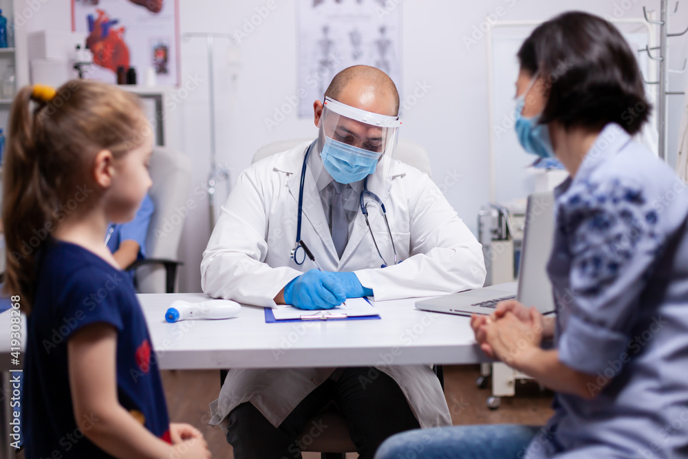 Pediatrician questioning patient about disease and writing answears in clipboard. Medical doctor in protective equipment against coronavirus discussing the symptoms in medical clinic Pediatrician questioning patient about disease and writing answears in clipboard. Medical doctor in protective equipment against coronavirus discussing the symptoms in medical clinic