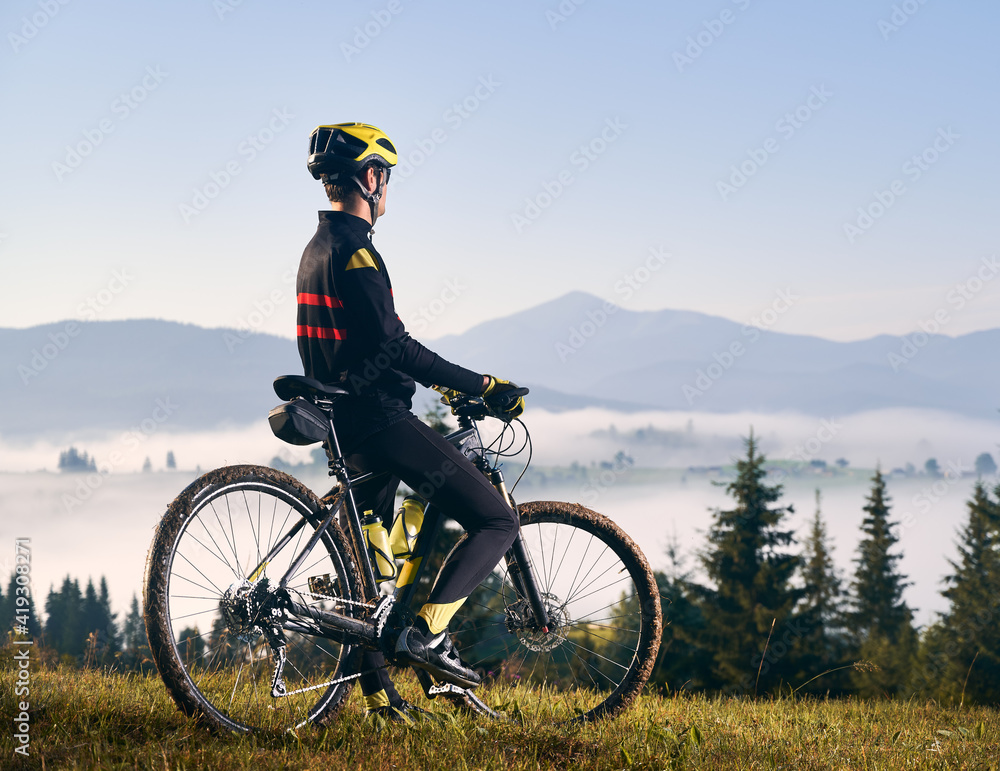 Obraz premium Back view of man in cycling suit standing with bicycle on grassy hill and looking at beautiful misty mountains. Male bicyclist in safety helmet enjoying view of majestic mountains during bicycle ride.
