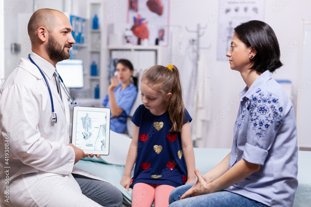 Pediatrician doctor explaining bones disease to mother using tablet with schelet image for sick child in hospital office. Physician specialist in pediatry medicine providing professional services Pediatrician doctor explaining bones disease to mother using tablet with schelet image for sick child in hospital office. Physician specialist in pediatry medicine providing professional services
