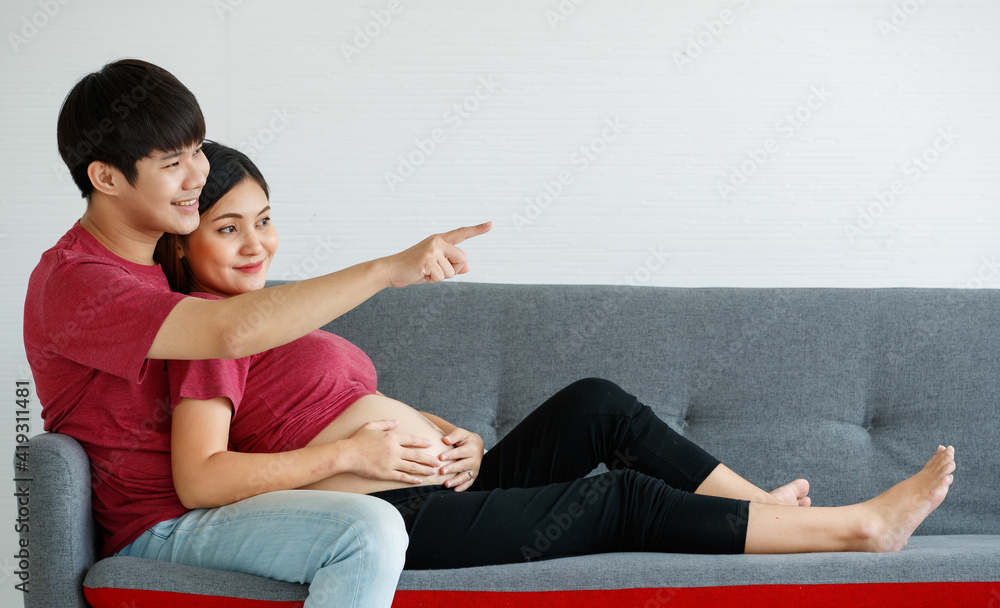 portrait of cute young Asian couple husband and wife sitting together on a couch at home. A man smiling and pointing something ahead while a pregnant woman leans on his chest happily