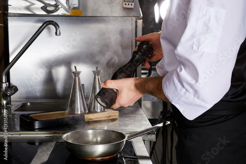 Chef add spices to dish cooking on fire, on stove. Cropped male cook in apron during preparing dish, in restaurant kitchen