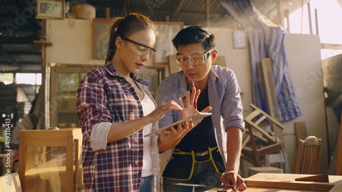 Asian male carpenter in safety goggles standing using a tape measure measuring the wooden door with Asian female carpenter standing next to him recording measurement results on the digital tablet.