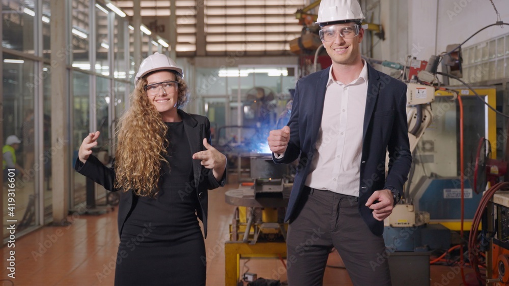 Two industrial engineers manager and secretary female wearing safety hardhat with goggle are celebrating business success and dancing having fun in factory.
