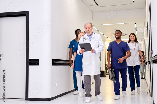 Group photo of young diverse medics during conversation with senior confident doctor in hospital corridor aisle. medicine concept