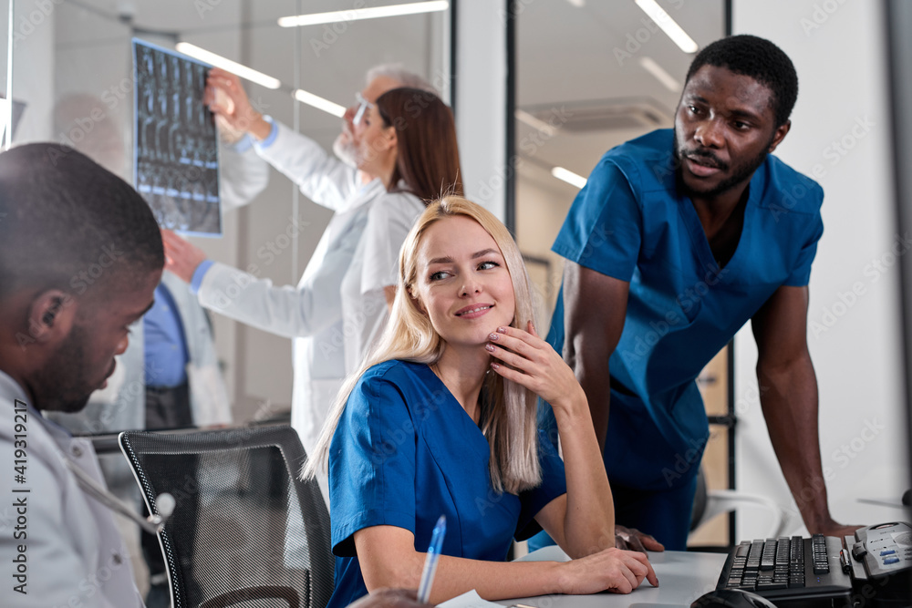 Obraz premium Interracial radiologists sitting at table in front of computer monitors with x-ray images and pleasantly discussing diagnosis in team at office. medicine concept