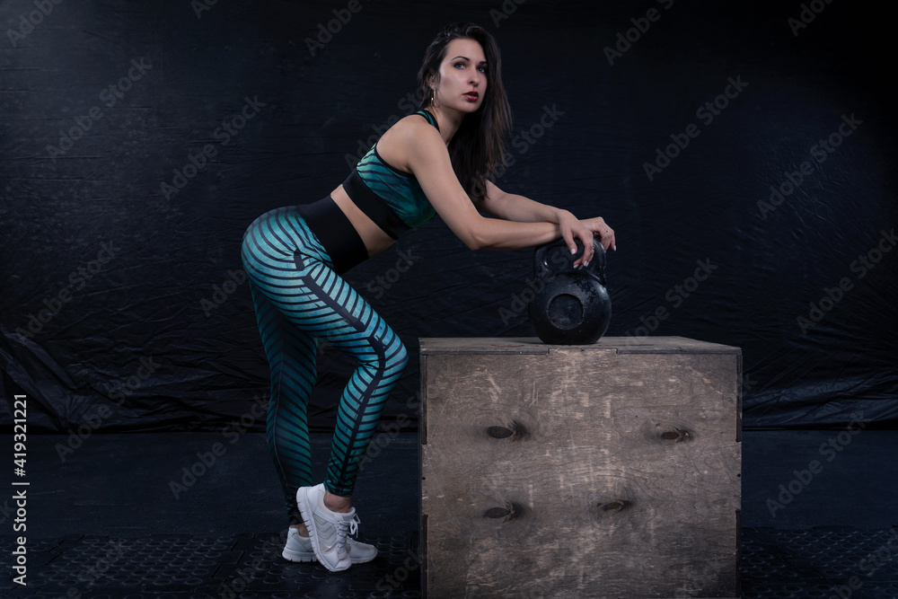 a girl in sportswear poses against a dark background, leaning against an iron weight that stands on a wooden box a girl in sportswear poses against a dark background, leaning against an iron weight that stands on a wooden box