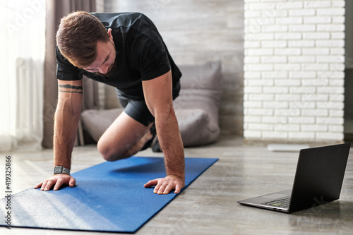 Fototapeta Naklejka Na Ścianę i Meble -  Athlete male doing exercises on mat watching online training on laptop, caucasian guy concentrated on sport and wellbeing, at home in living room