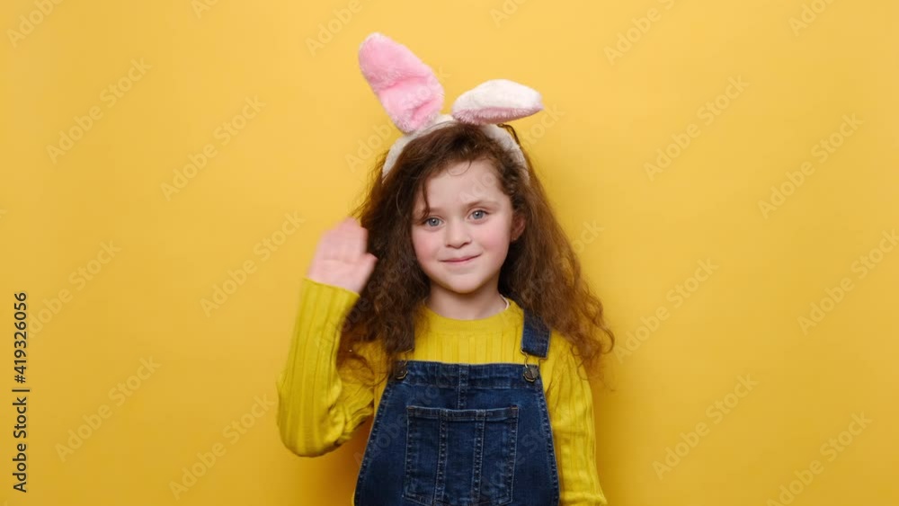 Joyful preschool little girl in bunny fluffy ears looking at camera with charming smile waving greeting with hand as notices someone, isolated on yellow background. Hello and Easter holiday concept Joyful preschool little girl in bunny fluffy ears looking at camera with charming smile waving greeting with hand as notices someone, isolated on yellow background. Hello and Easter holiday concept