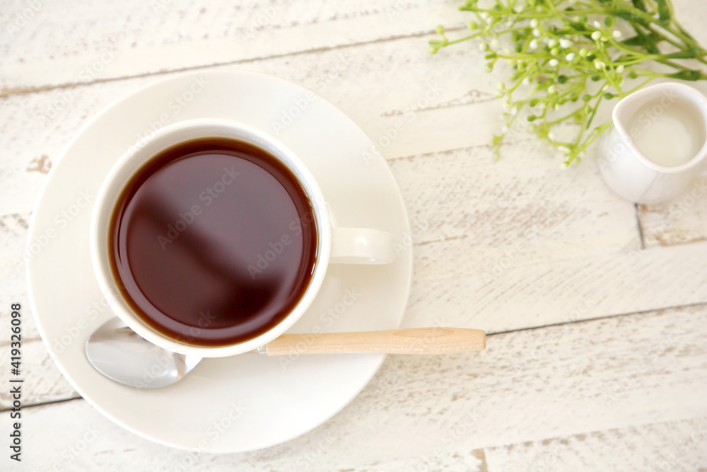 Hot coffee on a white wooden table, and a coffee spoon, a milk pitcher, and butterweed flowers. Hot coffee on a white wooden table, and a coffee spoon, a milk pitcher, and butterweed flowers.