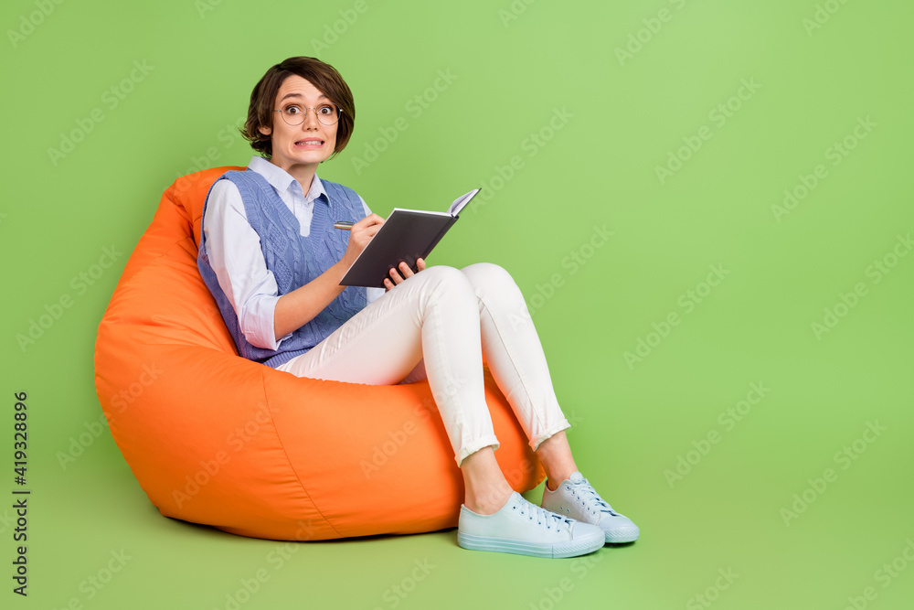 Portrait of attractive worried nervous girl sitting in bag chair writing notes plan to-do list isolated over green color background Portrait of attractive worried nervous girl sitting in bag chair writing notes plan to-do list isolated over green color background