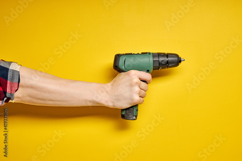 Green cordless battery powered drill on yellow background, cropped male hands holding tool for repair and building construction. copy space for advertisement