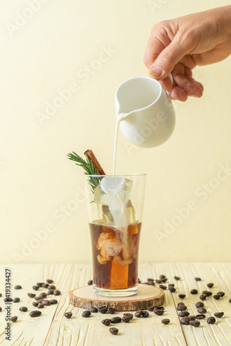 pouring milk in black coffee glass with ice cube, cinnamon and rosemary