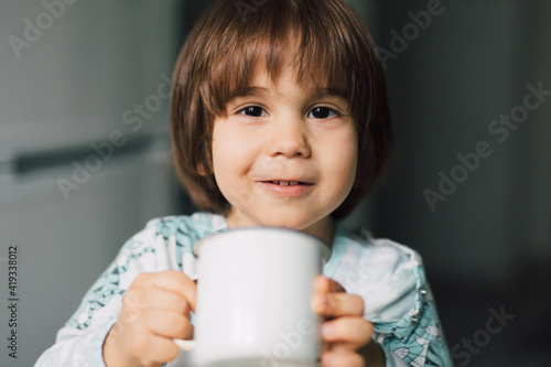 Cute smiling preschool boy holding a mug with milk at home. Kid with authentic smile, having breakfast.