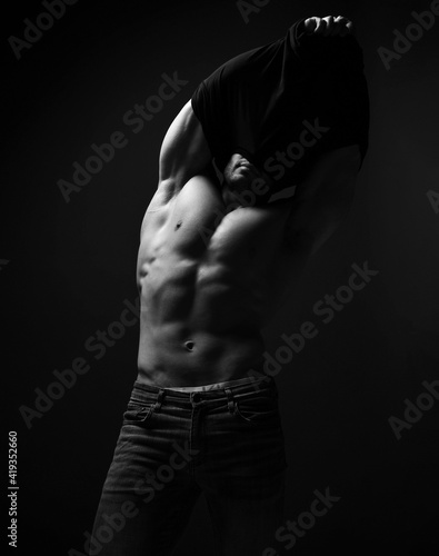 Strong muscular man, bodybuilder, weightlifter with perfect built athletic body stands taking off his shirt, getting undressed over dark background