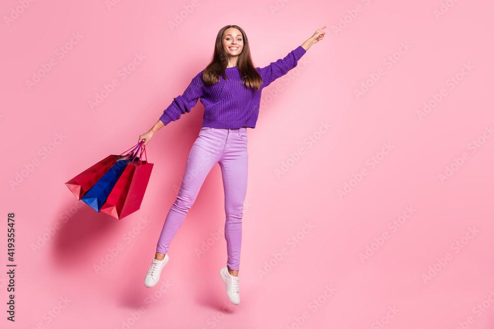 Full size photo of cheerful nice lady fly jumping hold bags direct finger empty space isolated on pink color background Full size photo of cheerful nice lady fly jumping hold bags direct finger empty space isolated on pink color background
