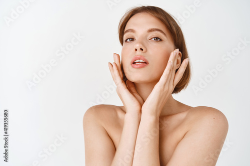 Beauty face. Young natural woman applying skin care facial cream, touching face and look at camera, standing on white background