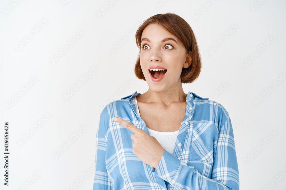 Excited girl pointing and looking at awesome promo deal, showing advertisement. Woman gasping and staring amazed at special offer, white background Excited girl pointing and looking at awesome promo deal, showing advertisement. Woman gasping and staring amazed at special offer, white background