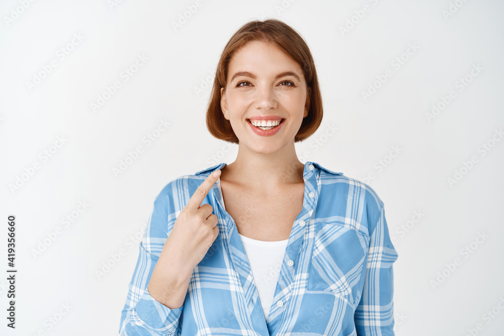 Portrait of smiling professional girl pointing at herself, self-promoting, talking about personal achievement, standing in casual clothes on white background Portrait of smiling professional girl pointing at herself, self-promoting, talking about personal achievement, standing in casual clothes on white background
