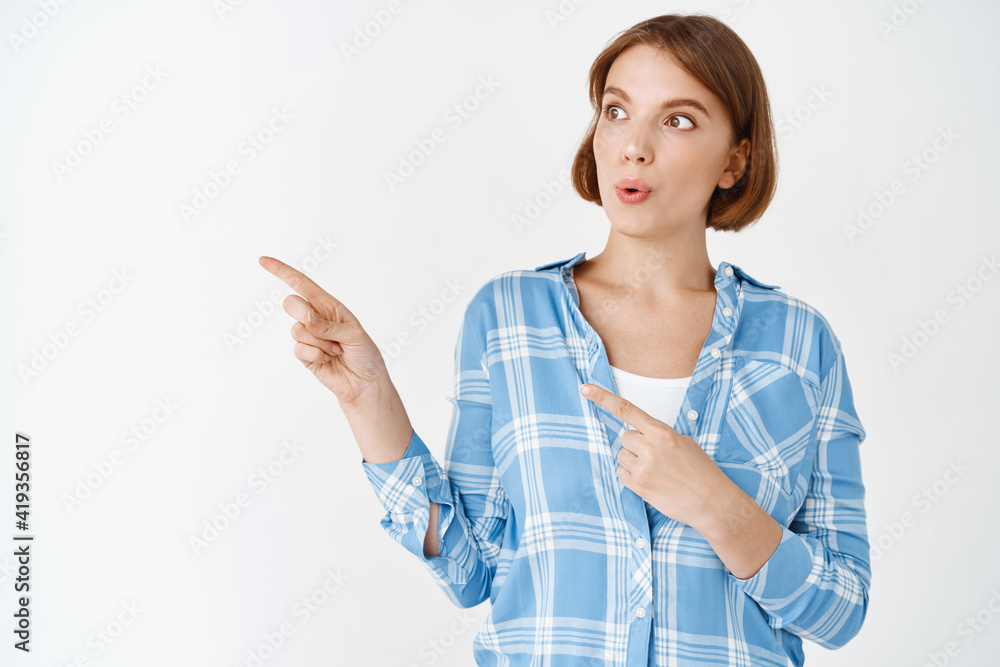 Wow, look. Excited cute girl with natural facial skin, whistle intrigued, pointing and gazing left at copy space, checking out advertisement, white background Wow, look. Excited cute girl with natural facial skin, whistle intrigued, pointing and gazing left at copy space, checking out advertisement, white background