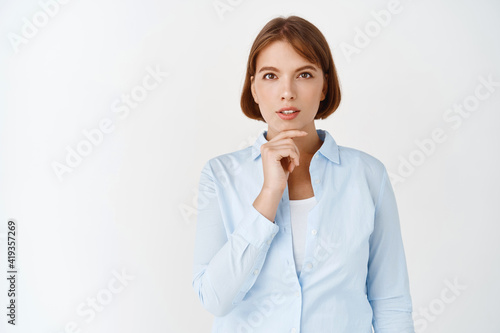 Getting inspiration. Portrait of young ceo female manager looking at camera thoughtful, have an idea, touching chin and pondering plan, standing on white background