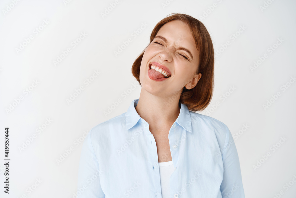 Portrait of carefree girl student smiling with eyes closed, showing tongue and having tongue, standing playful on white background Portrait of carefree girl student smiling with eyes closed, showing tongue and having tongue, standing playful on white background