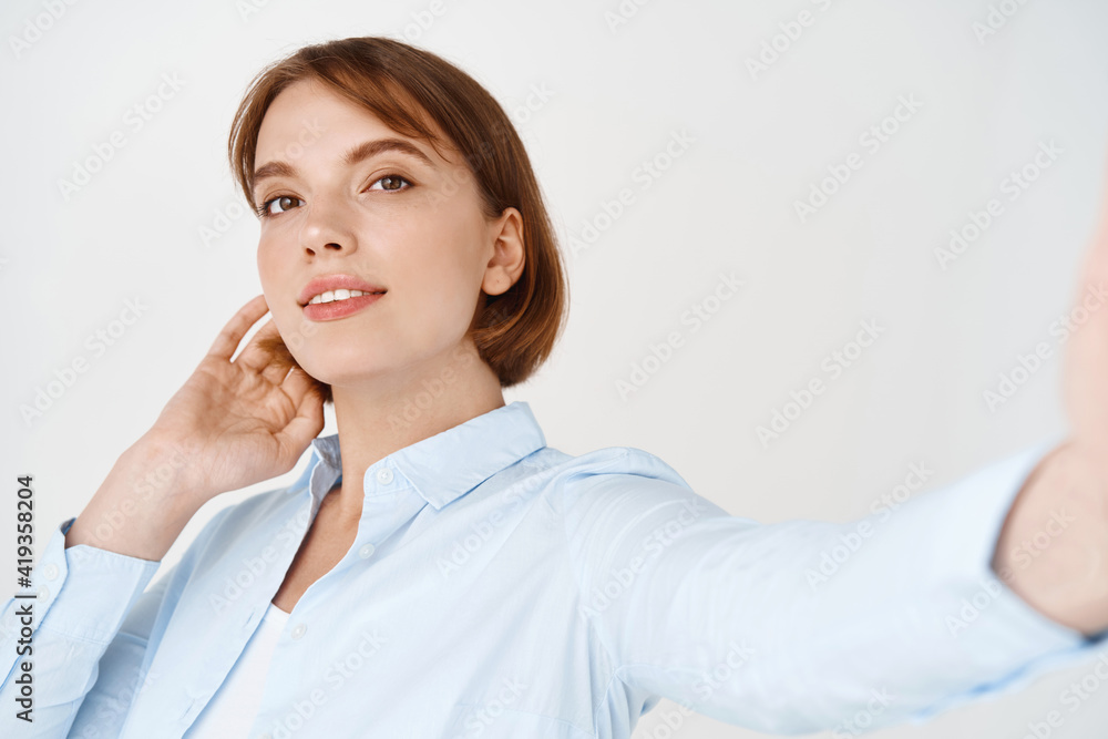 Beautiful candid woman in blouse, taking selfie on smartphone, holding phone in stretched out hand and smiling natural, standing on white background Beautiful candid woman in blouse, taking selfie on smartphone, holding phone in stretched out hand and smiling natural, standing on white background