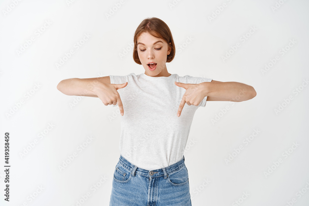 Excited and surprised young woman open mouth wondered, pointing and looking down amused, seeing interesting advertisement, standing against white background Excited and surprised young woman open mouth wondered, pointing and looking down amused, seeing interesting advertisement, standing against white background
