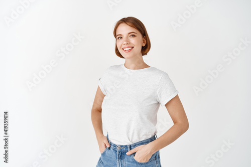 Cheerful natural girl in t-shirt and jeans smiling, looking upbeat, standing against white background © Cookie Studio