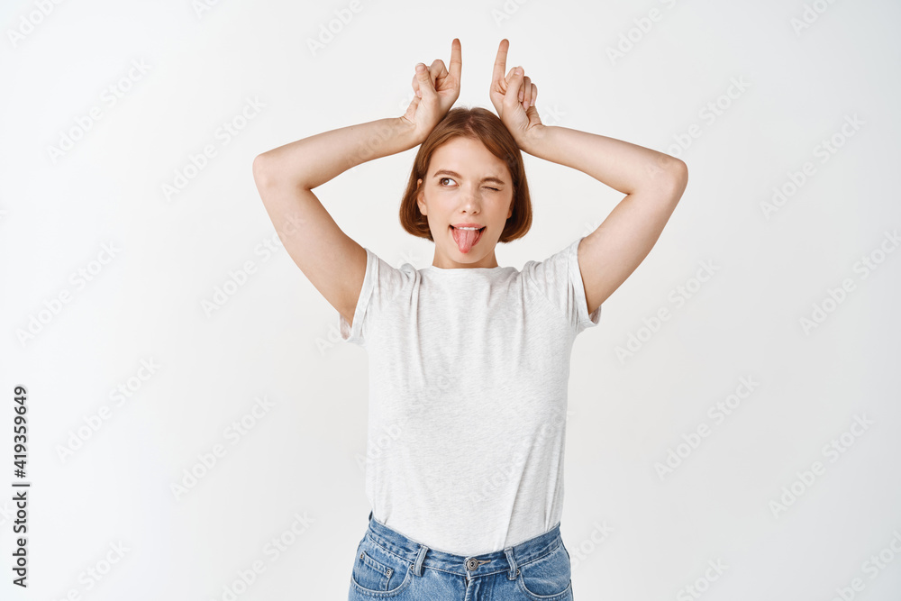 Happy young female model showing horns signs on head and tongue, looking aside with excited expression, standing on white background Happy young female model showing horns signs on head and tongue, looking aside with excited expression, standing on white background