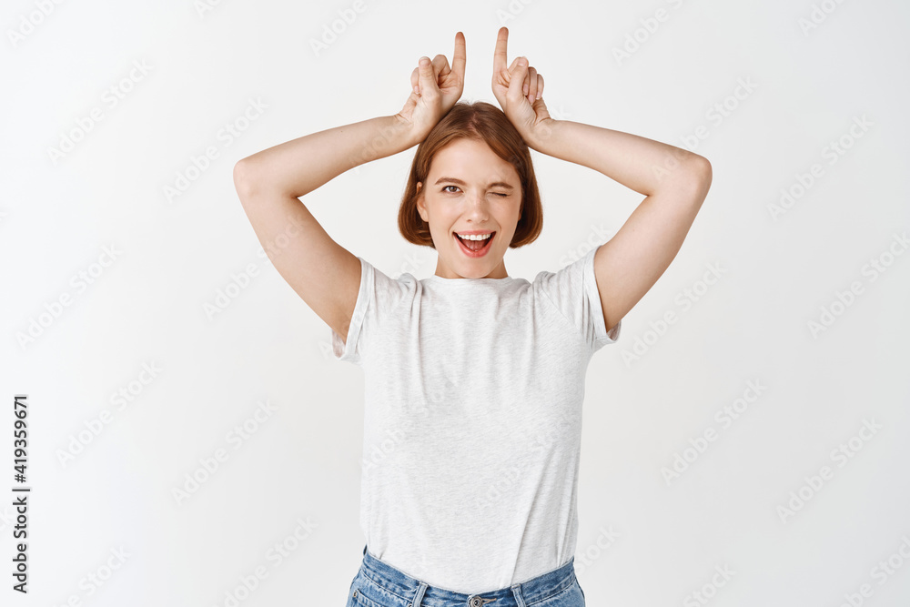 Cheerful young lady showing horns sign and winking at camera excited, standing upbeat in t-shirt and jeans, white background Cheerful young lady showing horns sign and winking at camera excited, standing upbeat in t-shirt and jeans, white background