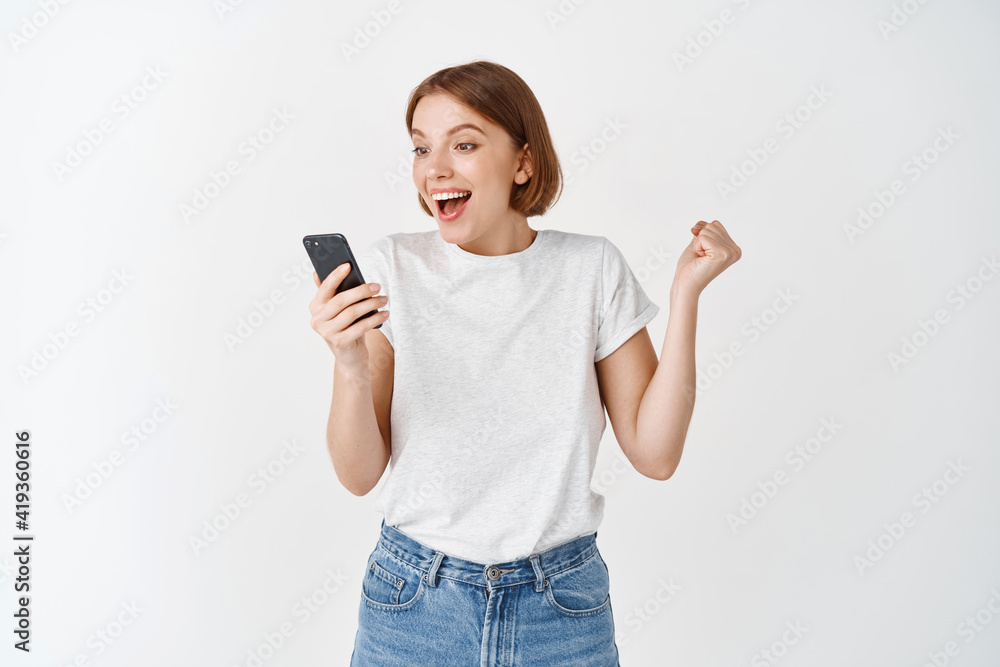 Happy woman winning on smartphone video game, raising hand up and cheering, shouting yes with joy, achieve online goal, standing on white background Happy woman winning on smartphone video game, raising hand up and cheering, shouting yes with joy, achieve online goal, standing on white background