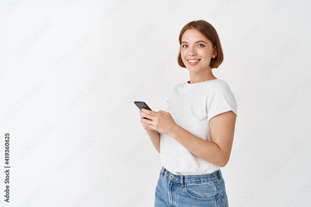 Young candid girl using smartphone and smiling at camera. Woman chatting on mobile phone app, standing against white background Young candid girl using smartphone and smiling at camera. Woman chatting on mobile phone app, standing against white background
