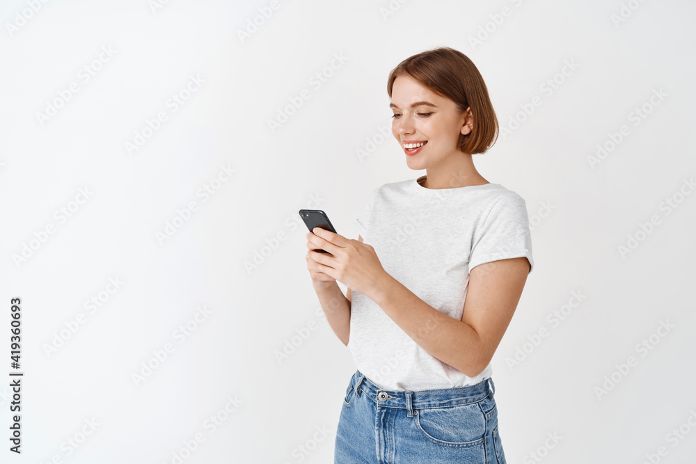 Portrait of smiling woman texting message, reading phone screen. Happy woman chatting on app, standing against white background Portrait of smiling woman texting message, reading phone screen. Happy woman chatting on app, standing against white background