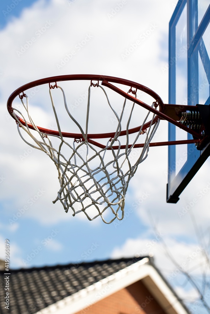 A portrait of an outdoor basketball rim with a net and a plexiglass backboard with blue lines on it in front of a cloudy sky. The basketball hoop or ring is orange and its pole is black.