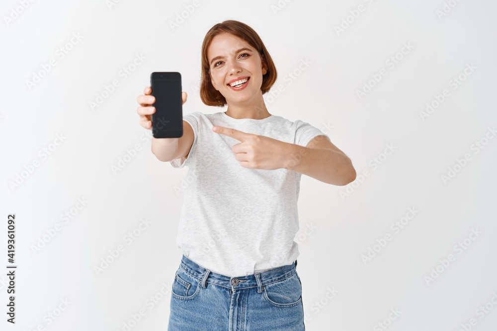 Check it out. Smiling beautiful woman in casual outfit, pointing at empty smartphone screen, showing online store or application, white background Check it out. Smiling beautiful woman in casual outfit, pointing at empty smartphone screen, showing online store or application, white background