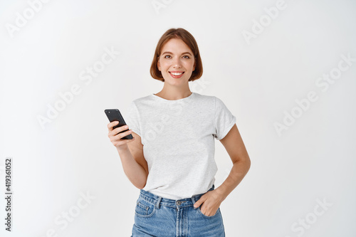 Portrait of smiling woman using smartphone, chatting on social media and looking at camera, standing with cell phone against white background