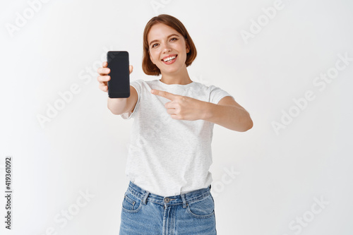 Check it out. Smiling beautiful woman in casual outfit, pointing at empty smartphone screen, showing online store or application, white background © Cookie Studio
