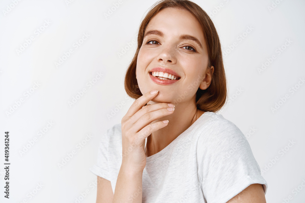 Close up of beautiful young woman with natural light make-up fresh face, touching chin and smiling tenderly, standing against white background Close up of beautiful young woman with natural light make-up fresh face, touching chin and smiling tenderly, standing against white background