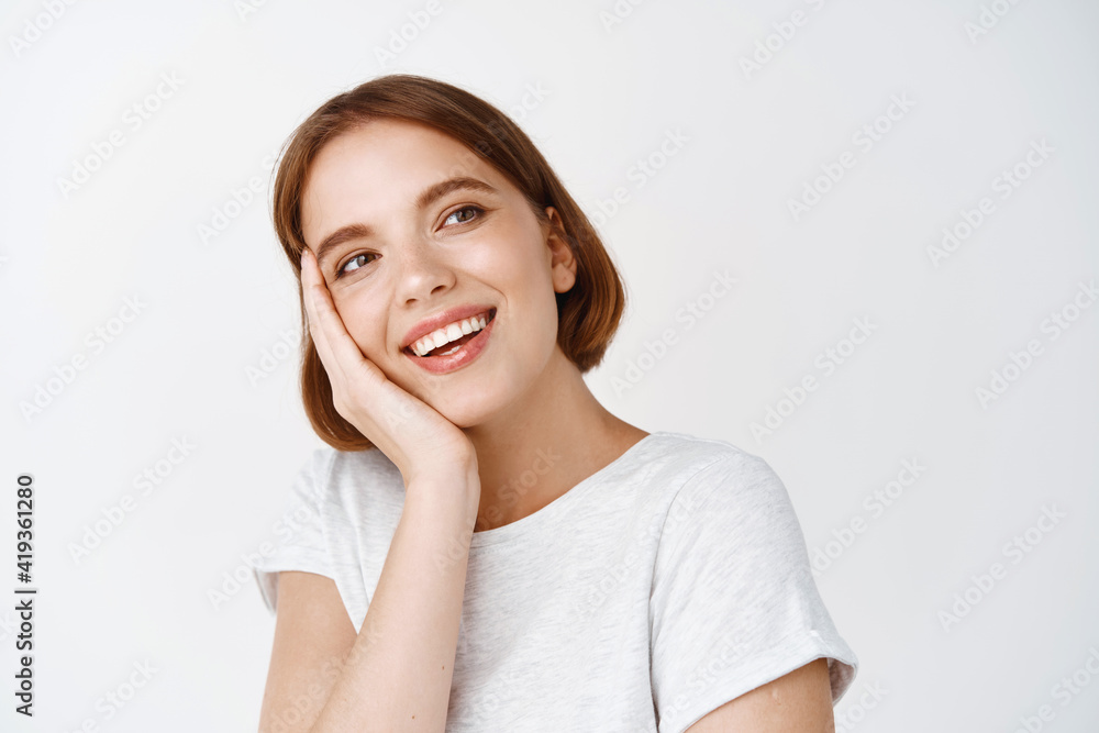 Beauty face. Tender happy woman with short hair, touching face and looking aside with dreamy smile, standing in t-shirt against white background Beauty face. Tender happy woman with short hair, touching face and looking aside with dreamy smile, standing in t-shirt against white background