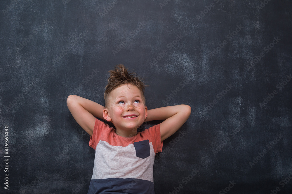 Smiling freckled kid boy dreaming. Child with hands behind head looking above. Boy against black school blackboard. Smiling freckled kid boy dreaming. Child with hands behind head looking above. Boy against black school blackboard.
